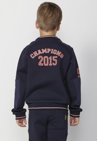 Sudadera azul marino con "CAMPEONES 2015" impreso en rojo en la parte posterior, con acentos texturizados en los hombros y puños acanalados.