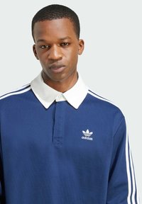Navy blue polo shirt with a white collar, featuring white three-stripe accents on the shoulders and a small white Adidas logo on the chest.