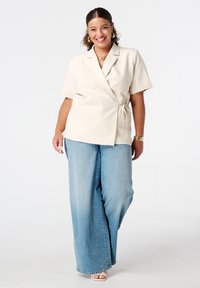 Blazer beige en style wrap avec des manches courtes, noué à la taille, associé à un jean ample taille haute bleu clair et des sandales à talons neutres.
