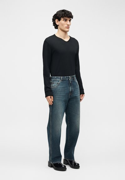 Νέος άνδρας που στέκεται, φοράει μαύρο μακρυμάνικο πουκάμισο, μπλε τζιν με ίσια μπατζάκια και μαύρα γυαλιστερά παπούτσια με πάτο, σε απλό φόντο.