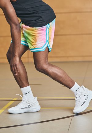 Person iført farverige shorts og hvide Nike-sneakers med matchende sokker, der bøjer knæ på en indendørs basketballbane.