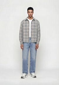 Άνδρας που στέκεται φορώντας γκρι καρό πουκάμισο με κουμπιά, άσπρο T-shirt, ανοιχτό μπλε τζιν και λευκά αθλητικά παπούτσια, μπροστά σε απλό φόντο.