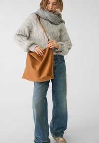 Donna che indossa un maglione grigio a maglia grossa e una sciarpa, jeans a gamba larga blu, sneakers beige, mentre tiene una grande borsa a tracolla in pelle marrone.