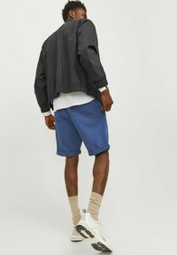 Veste noire courte avec des poignets élastiques, associée à un short bleu retroussé, des chaussettes beiges et des baskets blanches. Fond neutre.