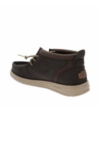 Scarpa slip-on marrone scuro con punta arrotondata, suola leggera beige e lacci decorativi. Presenta una patch con logo sul tallone.