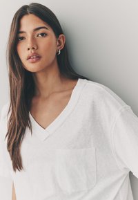 White V-neck t-shirt made from lightweight fabric, featuring a front pocket and relaxed fit. Smooth texture and subtle stitching details.