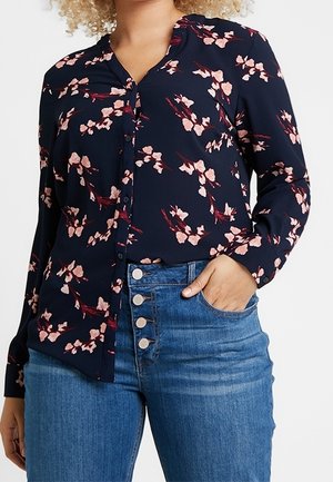 Mujer con blusa azul marino con estampado floral rosa y vaqueros azules de talle alto con cuatro botones metálicos visibles.