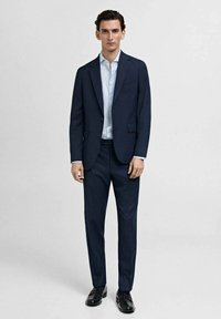 Costum de marin cu un sacou pe măsură și pantaloni asortați. Cămașă ușor albastră, proaspătă, dedesubt, completată de pantofi din piele neagră.
