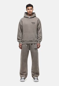 Kapuzensweatshirt und passende Jogginghose in einem gedämpften Grauton. Der Stoff erscheint weich und hat eine lockere Passform. Auf der Vorderseite sind aufgedruckte Logos zu sehen.