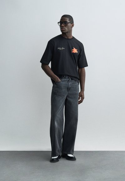 Άνδρας που φοράει υπερμεγέθη μαύρο T-shirt με γραφικό πουλιού, φαρδιά μαύρα τζιν, μαύρα γυαλιά και μαύρα παπούτσια με λευκές κάλτσες, στέκεται στο εσωτερικό.