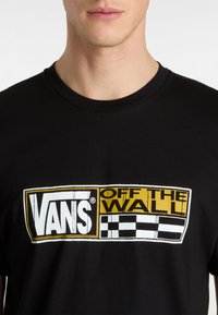 Svart bomullströja med en stor Vans-logotyp i gult, vitt och svart samt ett rutmönster under texten "OFF THE WALL."