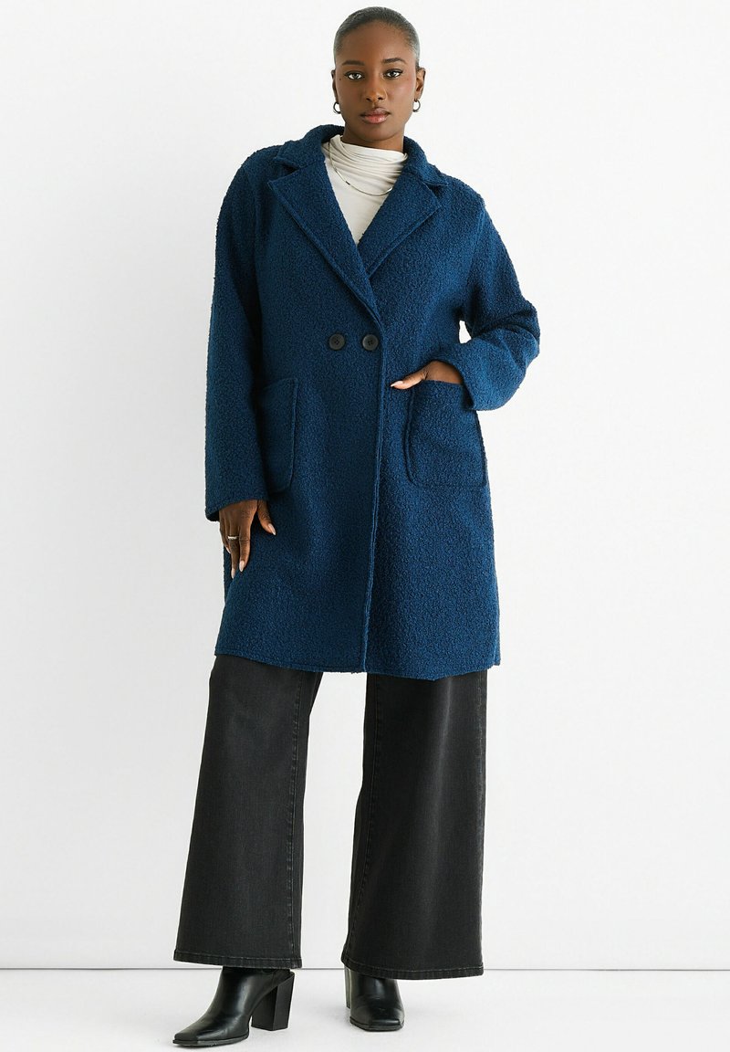 Navy blue textured coat with a tailored fit, lapel collar, two front pockets, and double button closure, paired with black wide-leg pants.