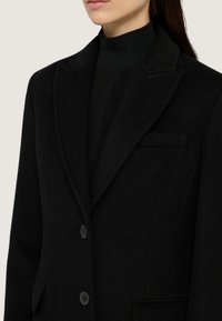 Černý kabát ze směsi vlny s propracovaným designem, klopovými límci a dvěma knoflíky. Obsahuje jedno náprsní kapsu a boční kapsy.