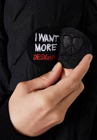 Černá prošívaná bunda s dvěma textilními nášivkami. Jedna nášivka je kruhová s vyšitým textem "CHCI VÍCE" v bílé a červené barvě, druhá má symbol míru.