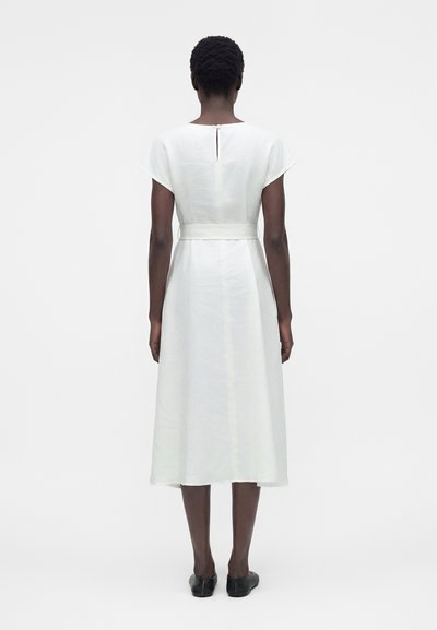 Γυναίκα που στέκεται με την πλάτη, φοράει λευκό μίντι φόρεμα με κοντά μανίκια και ζώνη, καθώς και μαύρα επίπεδα παπούτσια πάνω σε μονόχρωμο λευκό φόντο.