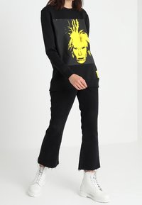 Černá mikina se žlutým grafickým motivem, doplněná černými rozšířenými kalhotami a bílými robustními botami. Hladká látka, volný střih, minimalistický styl.