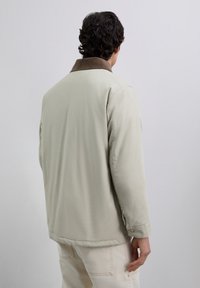 Man with dark curly hair wearing a light beige jacket with a brown collar, seen from the back against a plain gray background.