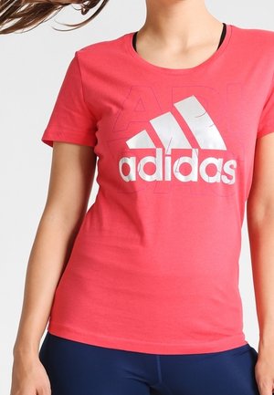 Kobieta w jasnoróżowej koszulce Adidas z dużym białym logo, w połączeniu z ciemnoniebieskimi legginsami, na jednolitym jasnym tle.