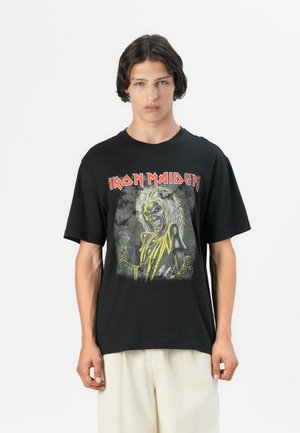Mustan puuvillateepaidan etäkulma, jossa on graafinen printti luukuvasta, kirkkaat keltaiset yksityiskohdat ja "IRON MAIDEN" -teksti rohkealla punaisella. Lyhyet hihat.