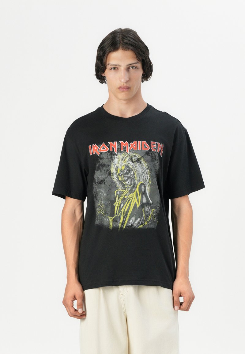 Melnas kokvilnas t-krekls ar grafisku zīmējumu par skeleta figūru, spilgti dzelteniem akcentiem un "IRON MAIDEN" tekstu treknrakstā sarkanā krāsā. Īsas piedurknes.