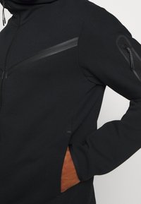 Svart hoodie med mjuk textur, utrustad med dragkedja fram, sidofickor och en kontrasterande brun detalj nära midjan.