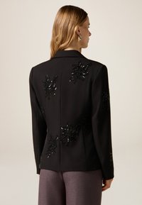 Zwarte blazer met bloemenapplicaties van pailletten op de achterkant, een getailleerde pasvorm en gestructureerde revers. Gecombineerd met grijze broek.