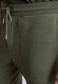Olijfgroene trainingsbroek van textuurstof, met een elastische tailleband en trekkoord, en zijzakken. Subtiele logodetail op het been.