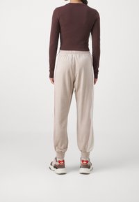 Žena viděná zezadu, oblékající béžové tepláky s růžovými bočními pruhy, přiléhavé tmavě hnědé tričko s dlouhým rukávem a béžové tenisky s červeným logem.