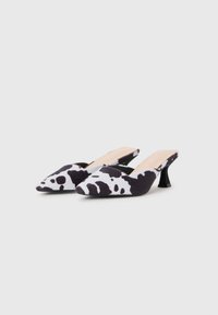 Mules à motif vache noir et blanc avec un bout pointu, un dessus en tissu doux et un talon bas et courbé. Présente un design à enfiler.