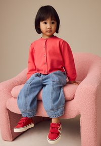 Jong kind met kort zwart haar zit op een gestructureerde roze stoel, draagt een rode cardigan, losse blauwe spijkerbroek en rode sneakers met witte zolen.