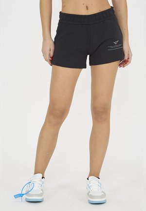 Kvinde iført sorte elastiske shorts i taljen og hvide sneakers med blå detaljer, stående mod ensfarvet baggrund.