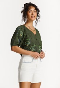 Blouse verte brodée avec un décolleté en V et des manches bouffantes, assortie à un short en denim blanc avec des coutures bleues sur les poches.