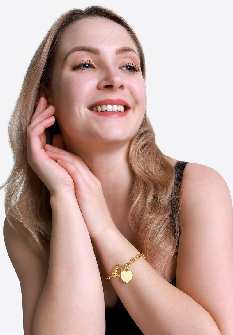 Bracciale con ciondolo a forma di cuore color oro, con design a catena, caratterizzato da una superficie liscia e una chiusura a girello, indossato su un polso con tono di pelle chiaro.