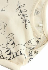Bodysuit beige avec un design imprimé noir, présentant des personnages de dessins animés et des plantes. Comprend des boutons-pression en bas. Texture de tissu douce.