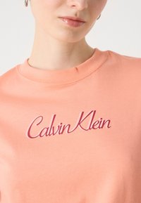 Persiko spalvos medvilninis marškinėliai su apvaliu iškirpimu, su raudonu užrašu švelniu šriftu "Calvin Klein" ant krūtinės. Paprasta ir lygi tekstūra.