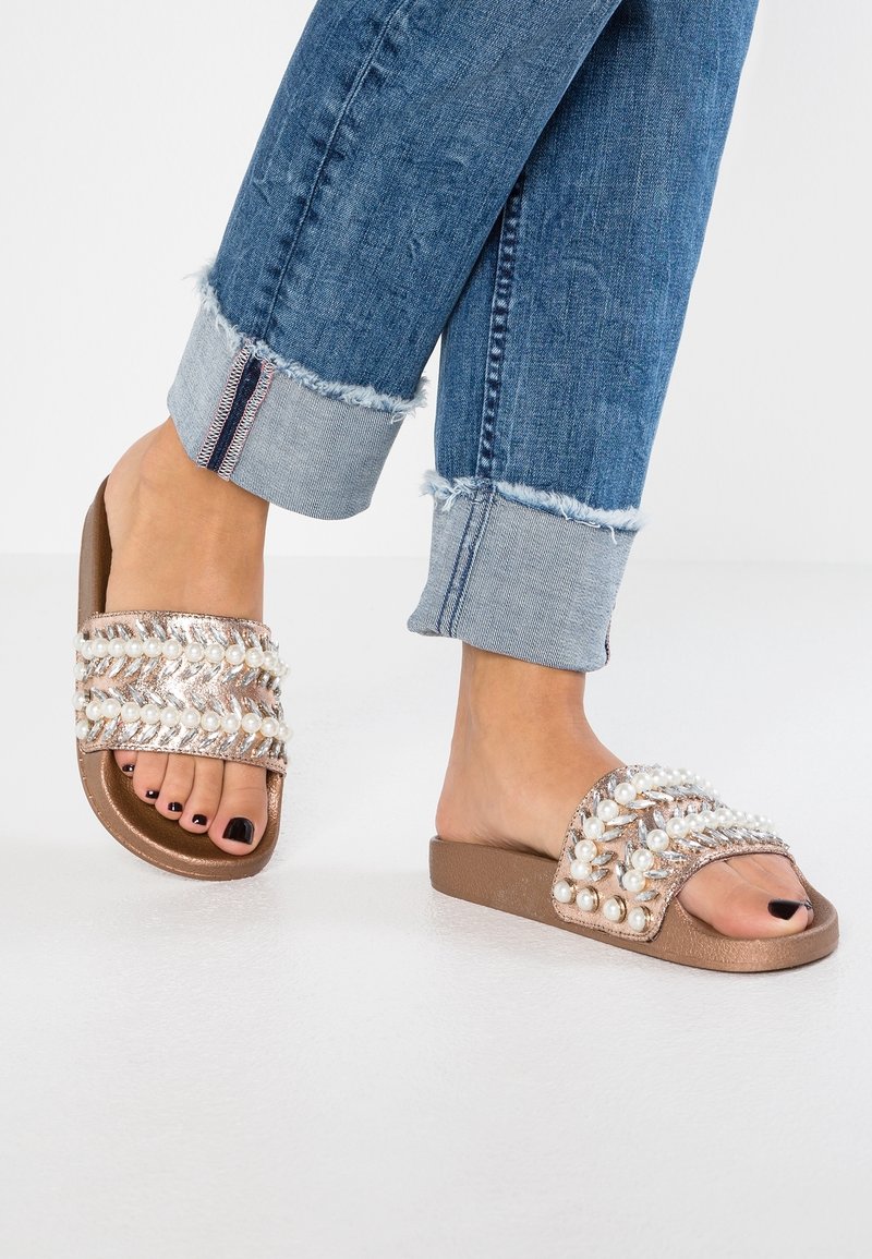 Des sandales à glisser avec une base marron, présentant un design tissé orné de perles blanches et d'accents métalliques. Portées avec un jean en denim retroussé.