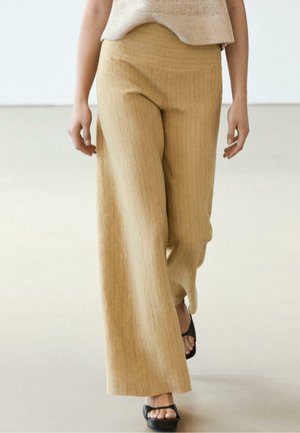 Persona che indossa pantaloni beige a gamba larga e sandali neri aperti sulle dita che cammina su un pavimento di colore neutro con uno sfondo semplice.