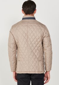 Beige quiltad jacka med diamantmönster och mörkrandig krage, med rak skärning och långa ärmar, sedd bakifrån.