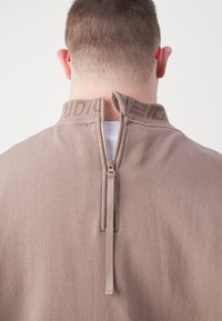 Uomo con capelli corti che indossa una maglietta beige con una cerniera parzialmente aperta sul collo sul retro, che mostra una t-shirt bianca.