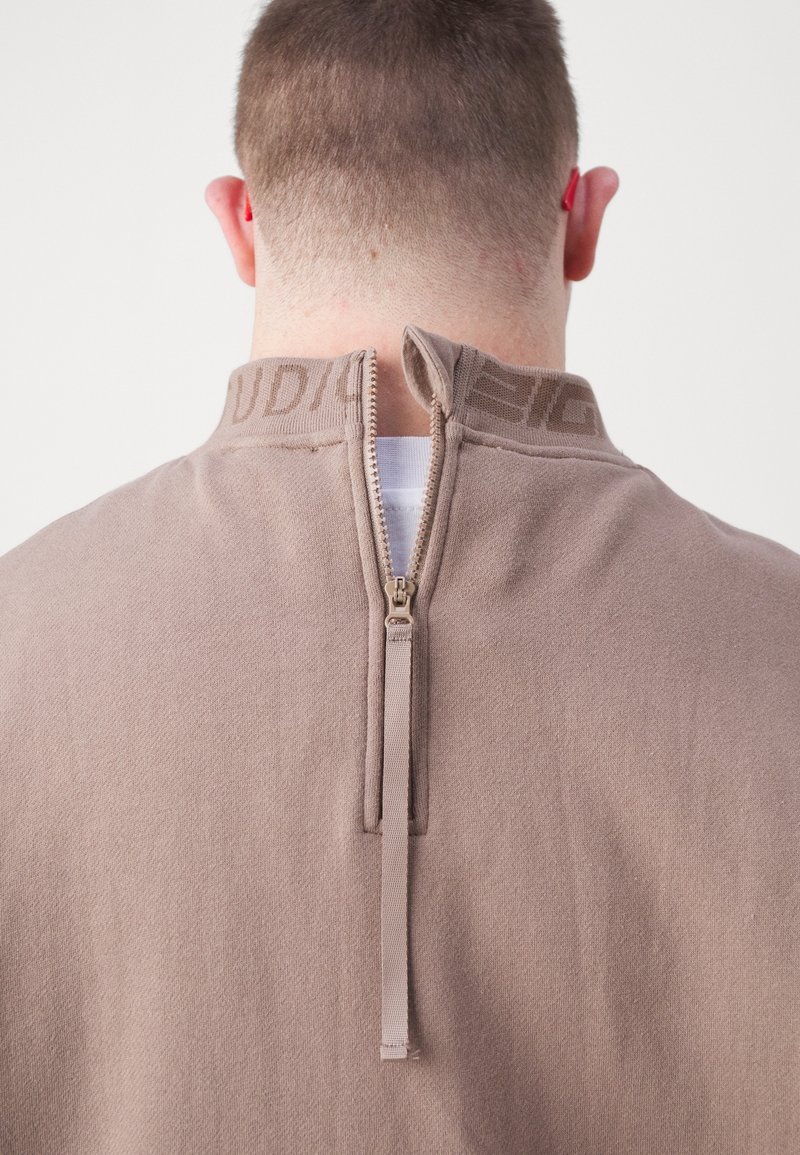 Uomo con capelli corti che indossa una maglietta beige con una cerniera parzialmente aperta sul collo sul retro, che mostra una t-shirt bianca.