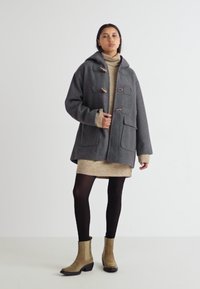 Manteau en mélange de laine gris avec capuche, fermetures à bascule et poches avant. Porté par-dessus une robe en tricot beige et des collants noirs, associé à des bottes chelsea beige.