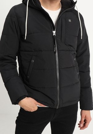 Homme portant une veste matelassée noire avec fermeture éclair frontale, poches latérales à boutons-pression, poche poitrine et cordons blancs à la capuche.