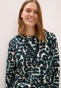 Sudadera con capucha que presenta un estampado animal abstracto multicolor en tonos verde azulado, negro y crema. Fabricada con una tela suave y texturizada, con detalles de cordón.