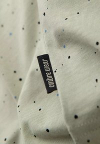 Lichtgrijze stof met een fijne textuur, versierd met verspreide zwarte en blauwe spikkels. Een zwart label met witte tekst is gedeeltelijk zichtbaar.