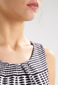 Hame dress tanpa lengan yang menampilkan kain berbentuk pola hitam dan putih, dengan leher bulat dan detail lipit halus di kerah.