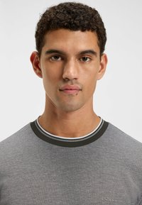 Grey short-sleeve t-shirt with textured fabric, contrasting black and white ribbed neckline, and small embroidered logo on the chest.