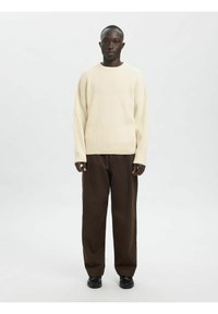 Svetlo bež rebrast pulover z ovratnim izrezom, kombiniran z ohlapnimi temno rjavimi hlačami in črnimi čevlji. Enostavna, teksturirana zasnova.