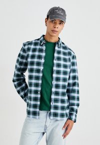 Groen en marineblauw geruit overhemd over een groen T-shirt, met lichtblauwe spijkerbroek en een grijze pet met witte tekst.