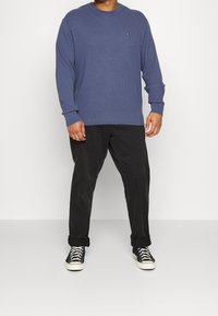 Niebieski prążkowany sweter z długim rękawem i okrągłym dekoltem, zestawiony z czarnymi spodniami i czarnymi trampkami. Sweter ma małe logo na piersi.