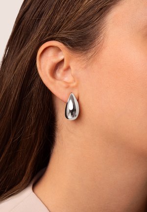 Oreille de femme portant une boucle d'oreille argentée lisse et brillante en forme de goutte, avec des cheveux bruns rabattus derrière l'oreille et un teint clair.
